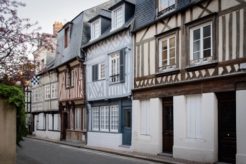 A charming street view displays a series of traditional half-timbered houses, showcasing varying colors and patterns. The architecture reflects historical design, with distinct window styles and intricate wooden beams. Lush greenery frames the scene, enhancing the overall atmosphere of the quaint neighborhood.
