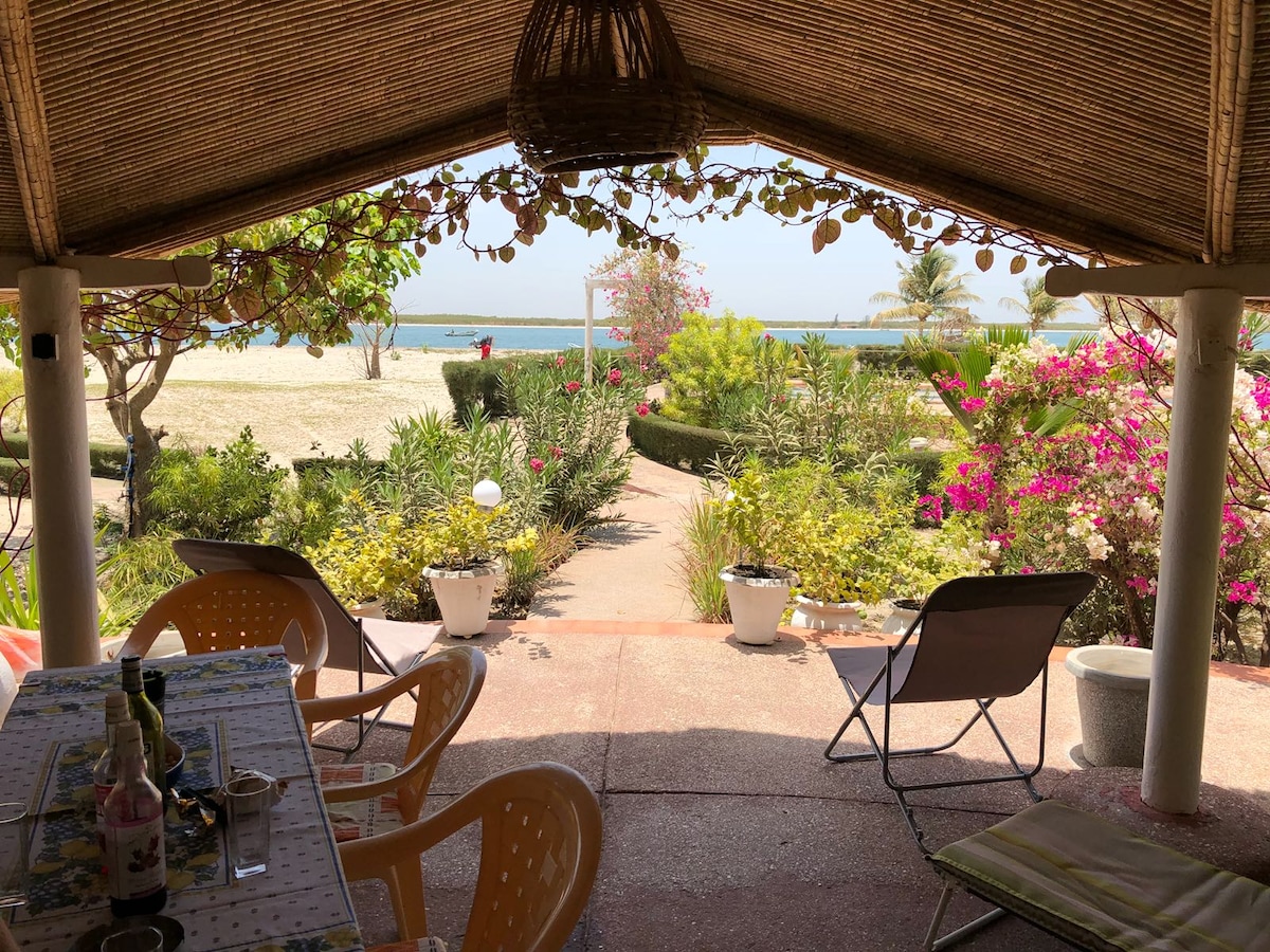 A covered terrace offers a view of a garden path lined with vibrant greenery and flowering plants, leading to a sandy beach. Two chairs are set on the terrace, inviting relaxation while enjoying the scenery of the beach and waters beyond.