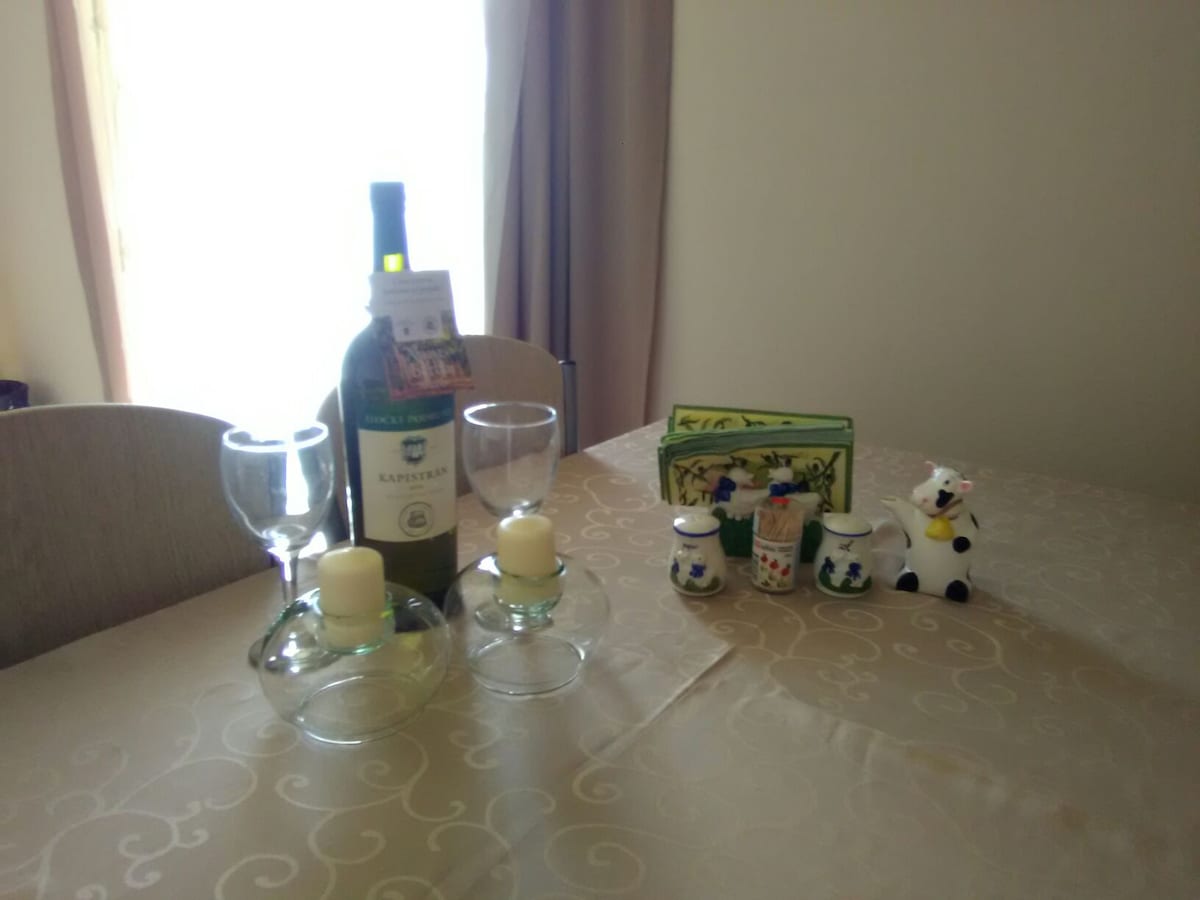 A dining table is set with decorative elements featuring a bottle of wine and two wine glasses. Small decorative candles are placed alongside ceramic milk containers and a playful cow figurine. The table is adorned with a neutral tablecloth, creating a cozy dining environment.
