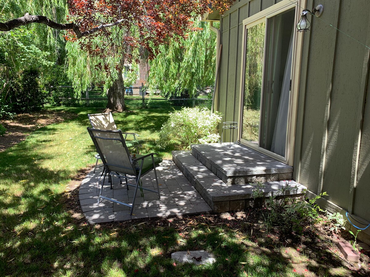 A small private patio features a stone surface with steps leading to a green entrance. A single chair is positioned for relaxation, surrounded by lush greenery and shaded by trees, creating a tranquil outdoor space.