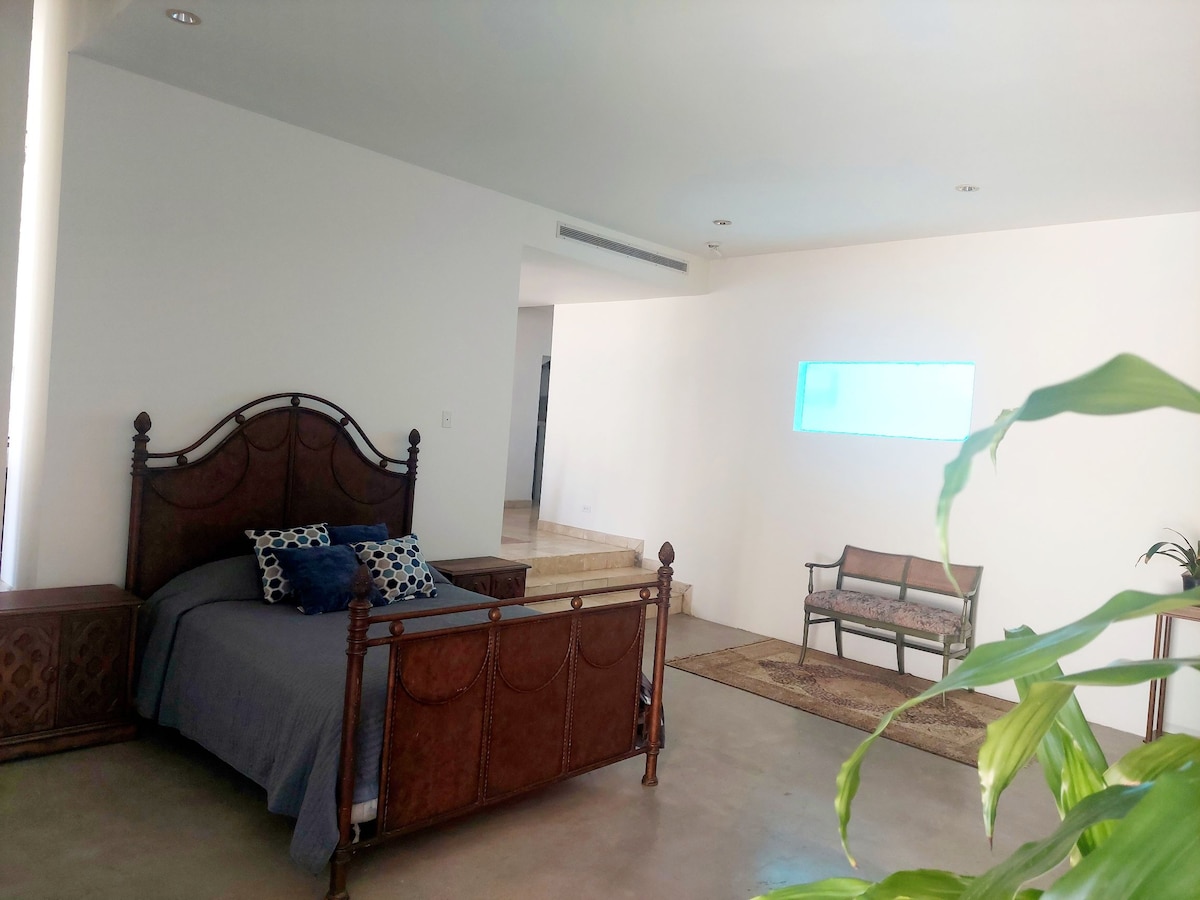The spacious room features a wooden bed framed with intricate carvings and adorned with decorative pillows. A comfortable seating area is visible across from the bed, complemented by a small rug. Natural light enters through a glass window, creating a soothing ambiance.