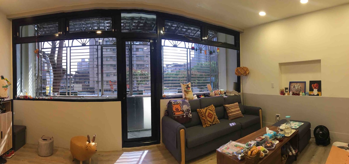 A light-filled living area features large windows with horizontal blinds, offering a view of the outside. A comfortable sofa adorned with decorative pillows faces a low coffee table surrounded by personal items. A small decorative animal is positioned on the floor.