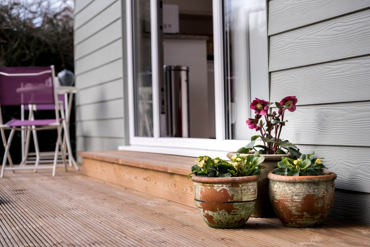 A small wooden terrace is accented by two terracotta pots containing blooming flowers. The entrance to the lodge is visible through expansive glass doors, while seating is provided by two light purple chairs positioned nearby.