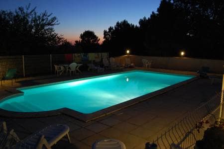 A spacious outdoor swimming pool is illuminated with soft turquoise light, surrounded by several lounge chairs. The area is bordered by fences and trees, creating a serene environment during twilight. The soothing glow of lights accentuates the pool's shape and contours.