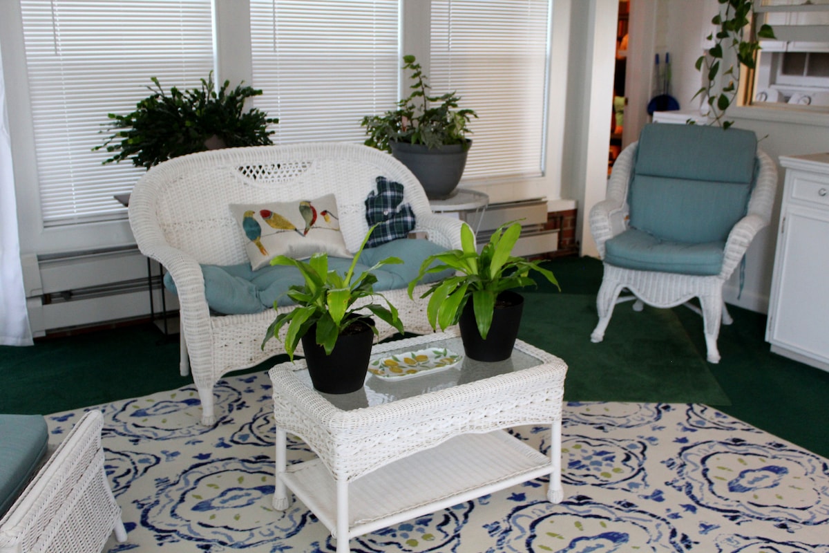 A sunroom features a white wicker sofa with blue cushions and a matching chair. A coffee table adorned with plants sits in front of the seating. The room is brightened by several large windows, allowing natural light to enhance the inviting ambiance.