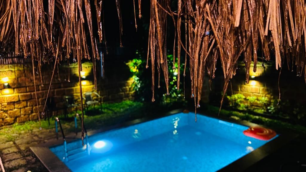A tranquil swimming pool is highlighted under soft illumination, surrounded by lush greenery. The pool area is framed by natural materials, with a thatched roof overhead. Soft water reflections shimmer across the pool's surface, enhancing the serene outdoor setting.