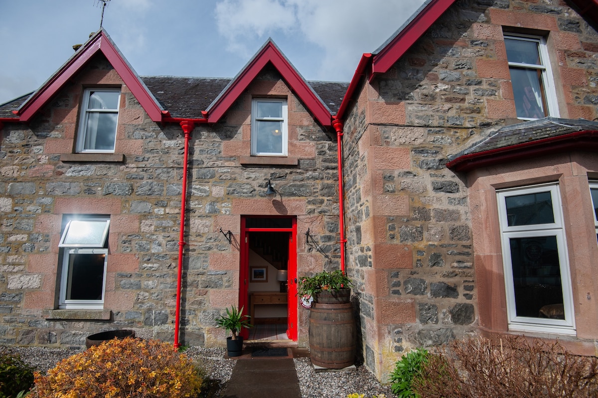 The exterior of a Victorian house is showcased, featuring a blend of stone and red accents. A welcoming entrance is highlighted by an inviting red door. Lush plants and a decorative barrel are positioned in the front garden, enhancing the charming façade.