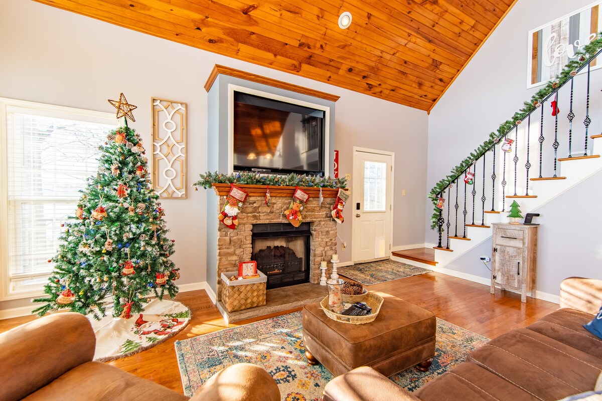 A spacious living area features a tall, vaulted ceiling with wooden beams. A decorated Christmas tree stands next to a stone fireplace, which is flanked by festive stockings. Opposite, stairs with a wooden railing lead to the upper floor, complemented by a decorative area rug.