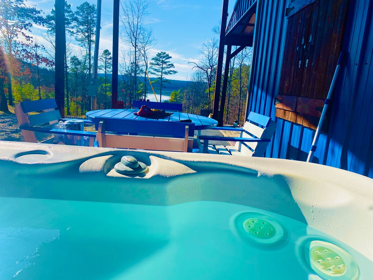 A hot tub is positioned at the forefront, with warm blue water visible. In the background, a wooden deck is featured alongside a round outdoor table and chairs. Trees frame the view, leading to a serene lake that reflects the clear sky.