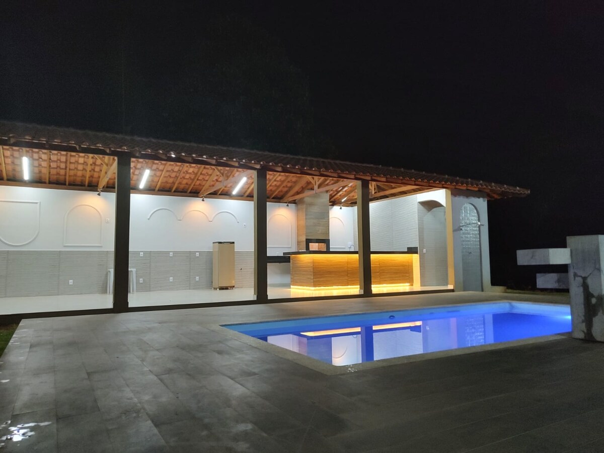 The image shows a spacious outdoor area featuring a well-lit gourmet space adjacent to a pool. The pool emits a soft blue glow, while warm lighting highlights the cooking area. The structure has a rustic roof supported by wooden beams, creating an inviting setting.