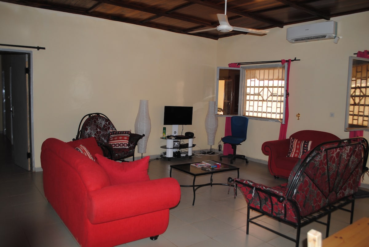 A spacious living area features two red sofas and a black coffee table. A television is situated on a stand, complemented by a modern chair. Natural light filters through two windows with decorative grilles, while a ceiling fan provides airflow.