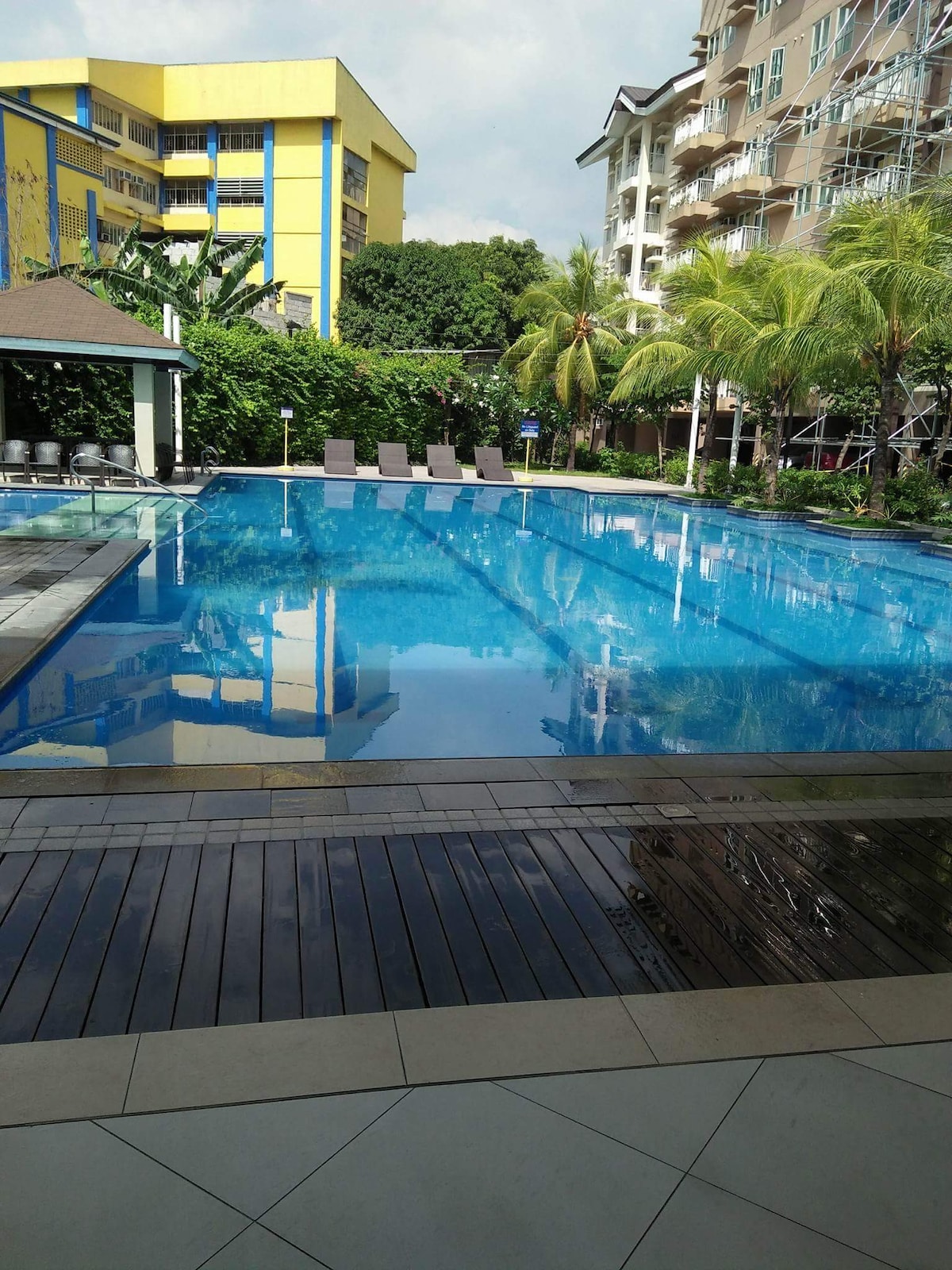 A clear swimming pool is surrounded by lush greenery and palm trees, reflecting the blue sky above. Lounge chairs are positioned along the poolside. Nearby, a shaded pavilion is visible, along with contemporary apartment buildings that frame the pool area.
