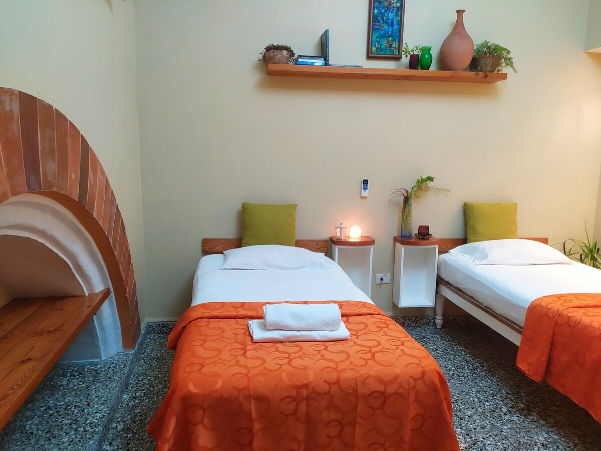 The room features two spacious beds with bright orange bedspreads, complemented by green cushions. A decorative shelf is placed above, displaying plants and art. A soft glow from candles adds warmth, while a cozy seating area with a wooden bench is present. Natural stone flooring is visible.