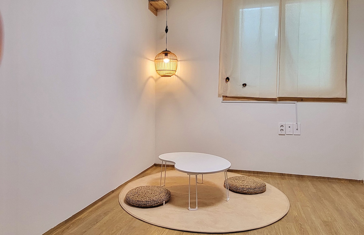 A quiet corner features a round cream mat on a wooden floor, with a low white table surrounded by two woven seating cushions. A pendant light provides soft illumination, while a simplified window with light curtains allows natural light to enter.