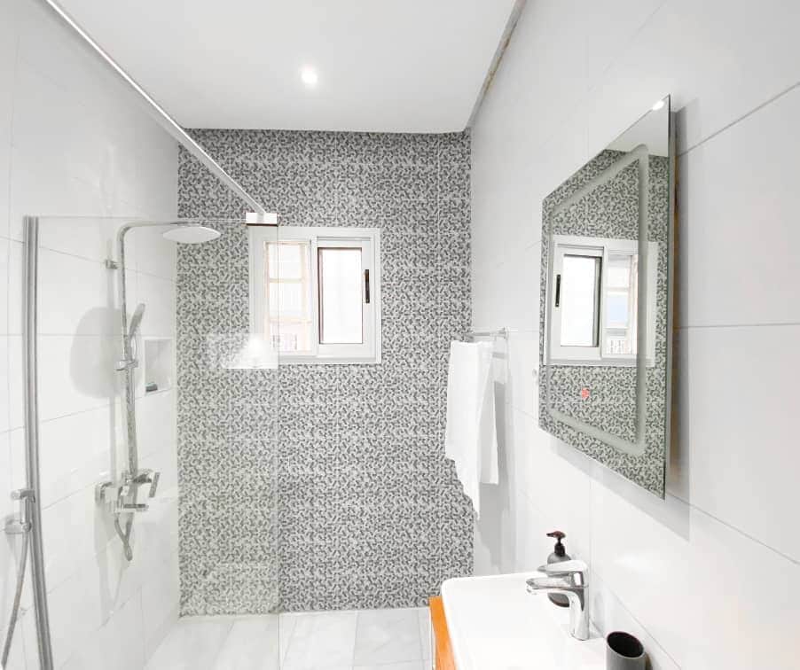 The modern bathroom features a walk-in shower with a glass partition and sleek fixtures. A stylish mirror hangs above the sink, which is accompanied by a towel rack. Natural light enters through a window adorned with subtle privacy grilles, complementing the contemporary tile design.