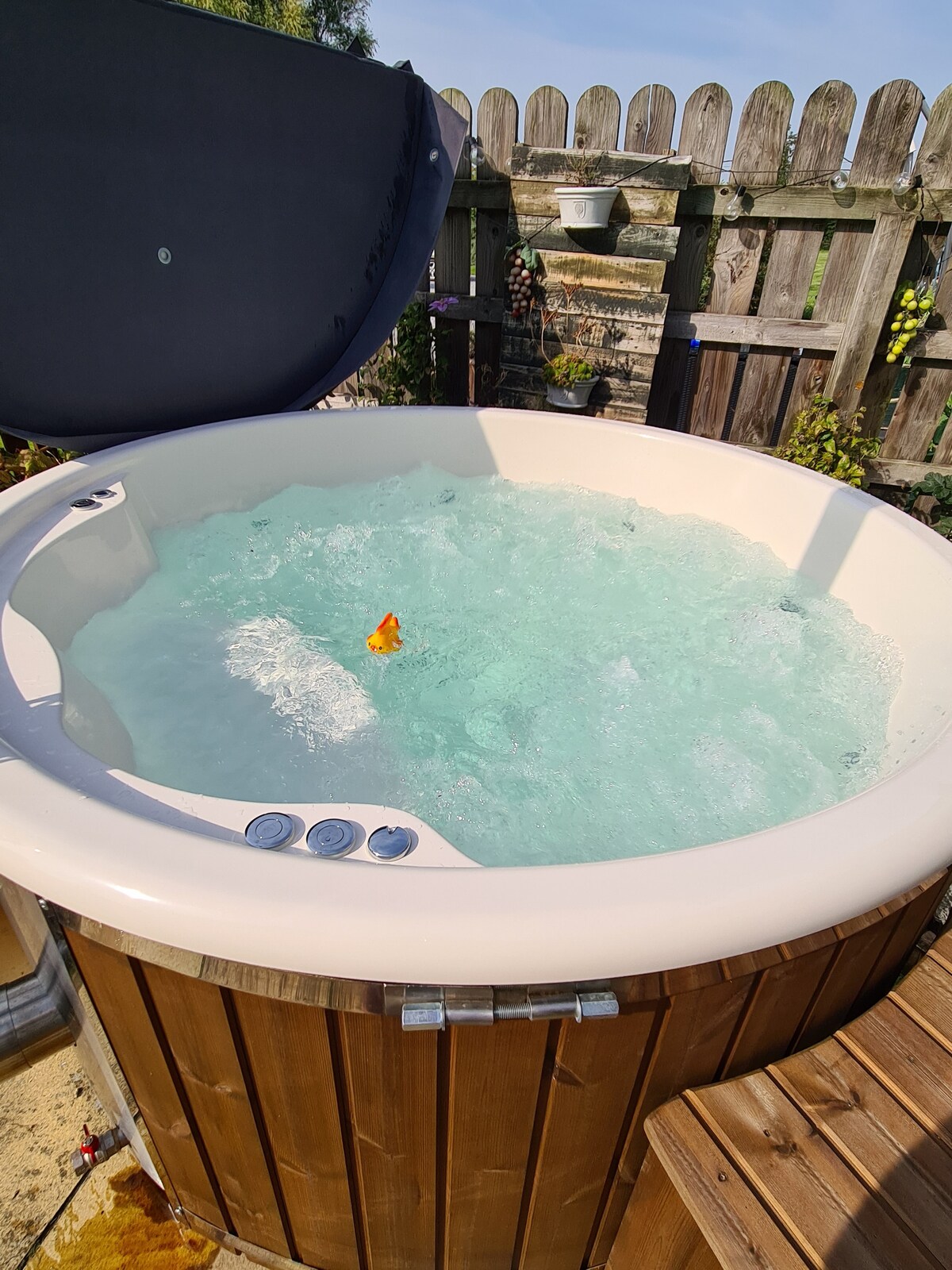A private wood-fired spa hot tub is featured, surrounded by wooden paneling. Clear water is seen bubbling gently, with a small rubber duck floating. The tub is set in a well-maintained outdoor area, with a wooden fence visible in the background.