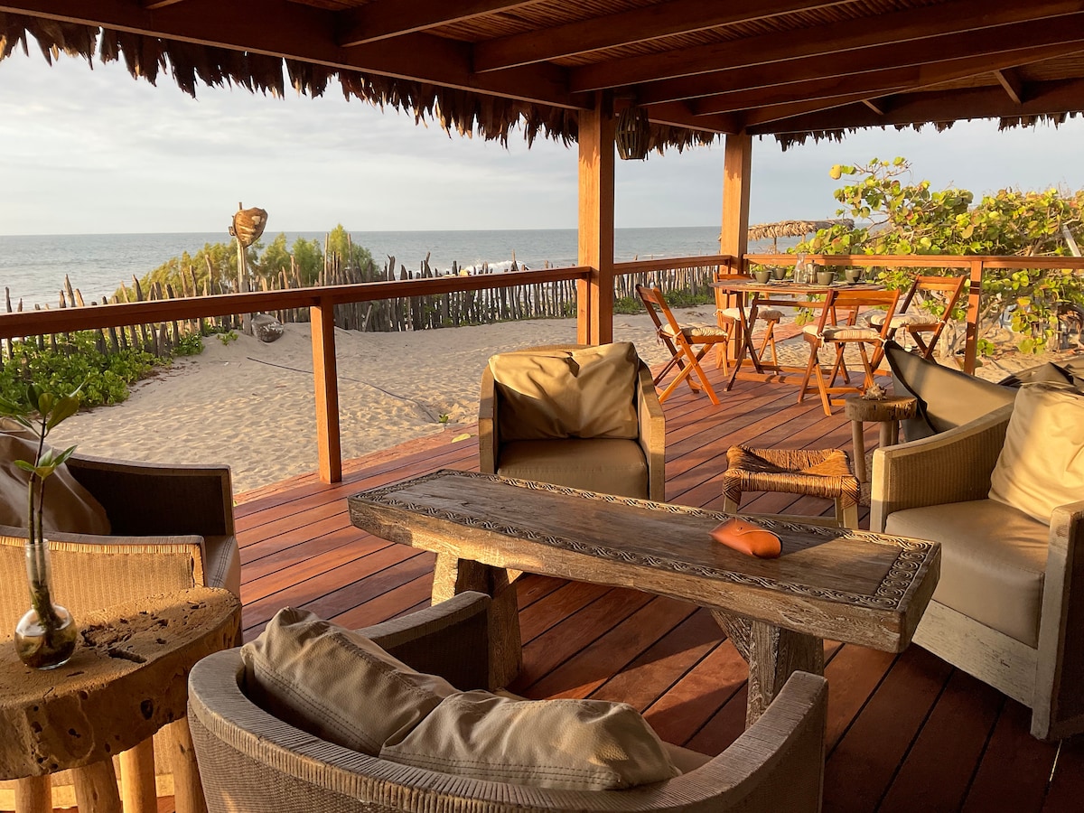A spacious deck features comfortable seating with wooden furniture, offering a clear view of the sandy beach and ocean. Lush greenery is visible nearby, enhancing the natural ambiance. The space is designed for relaxation, with soft sunlight illuminating the area.
