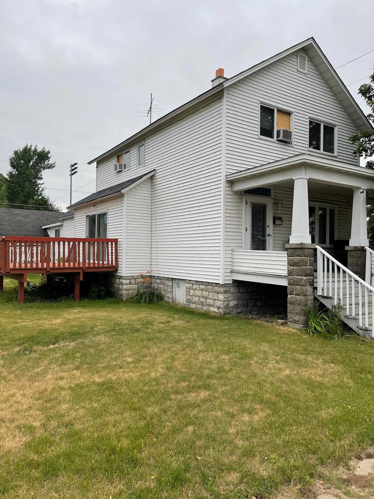 The exterior of the house displays a two-story structure with white siding and a covered front porch. A wooden deck with railings is visible, providing an inviting transition to the yard, which is framed by grass and shrubs.
