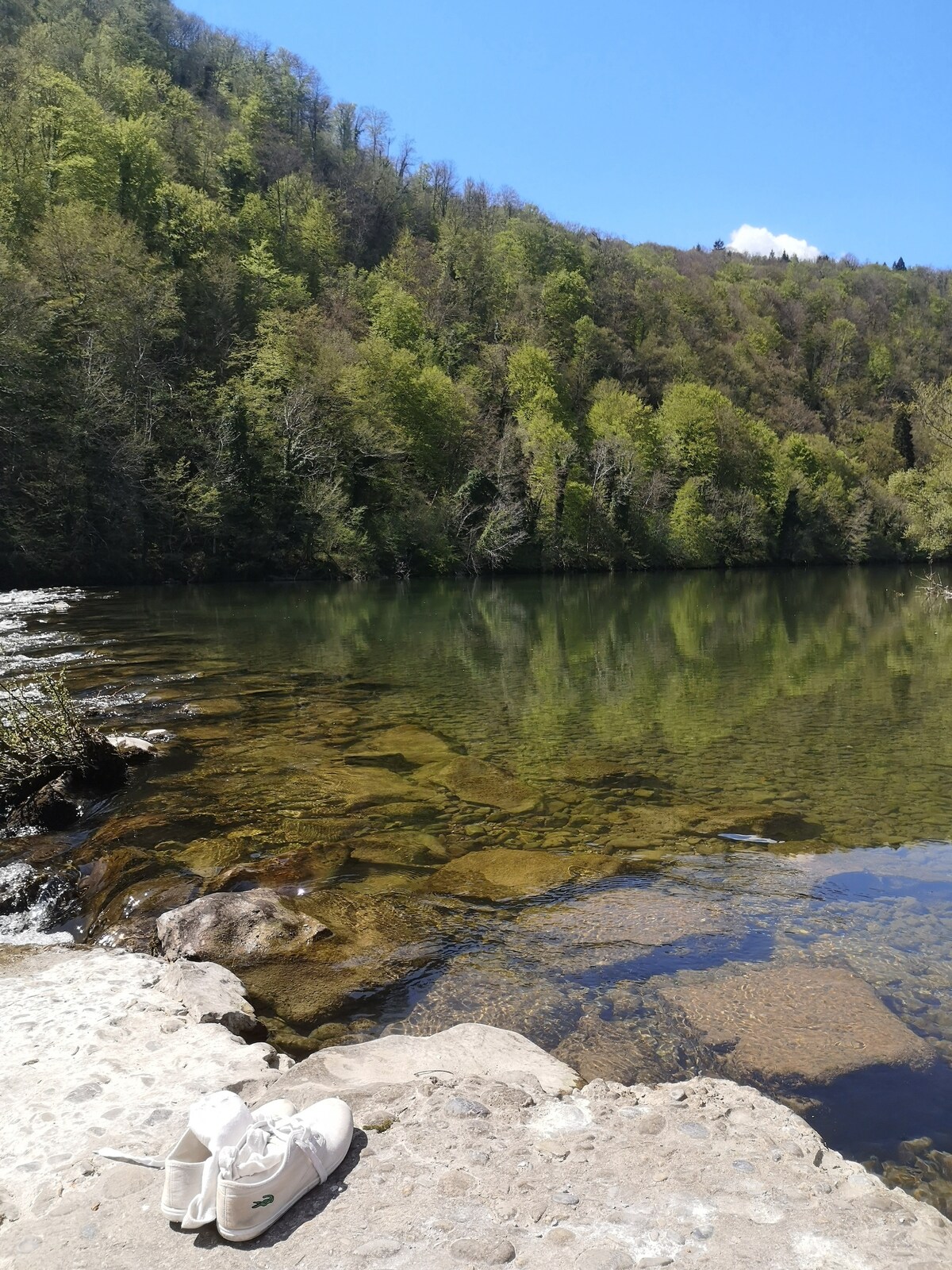 A calm riverbank is depicted with clear water reflecting lush green trees from the surrounding hills. Smooth stones are partially visible at the water's edge, while a pair of shoes rests on the sandy shore, suggesting a peaceful outdoor experience.