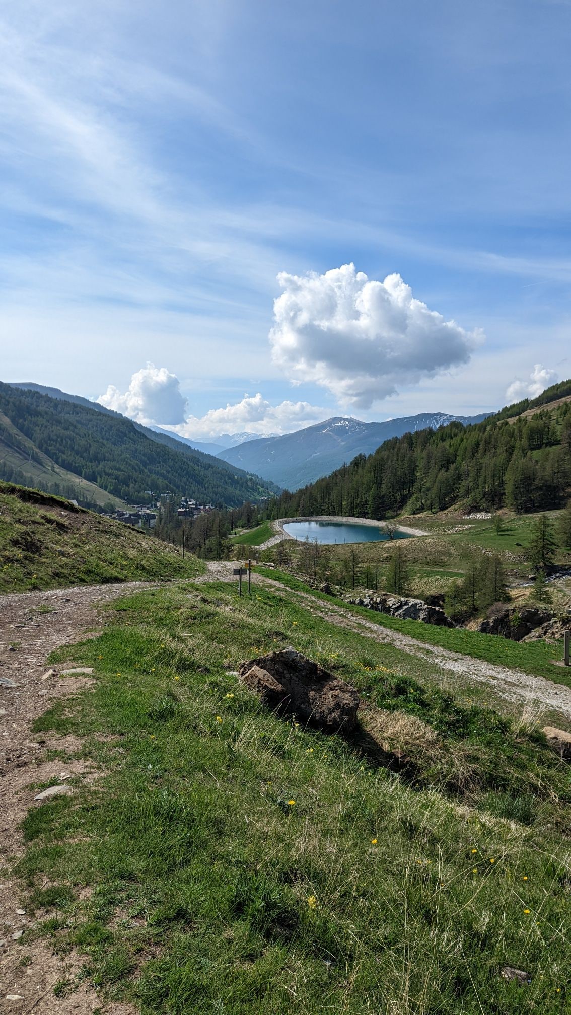 A scenic landscape showcases rolling hills and a serene lake nestled among mountains. The path in the foreground leads toward lush greenery, while distant peaks are partially shrouded in clouds. The expansive sky is filled with soft, fluffy clouds, creating a tranquil outdoor atmosphere.