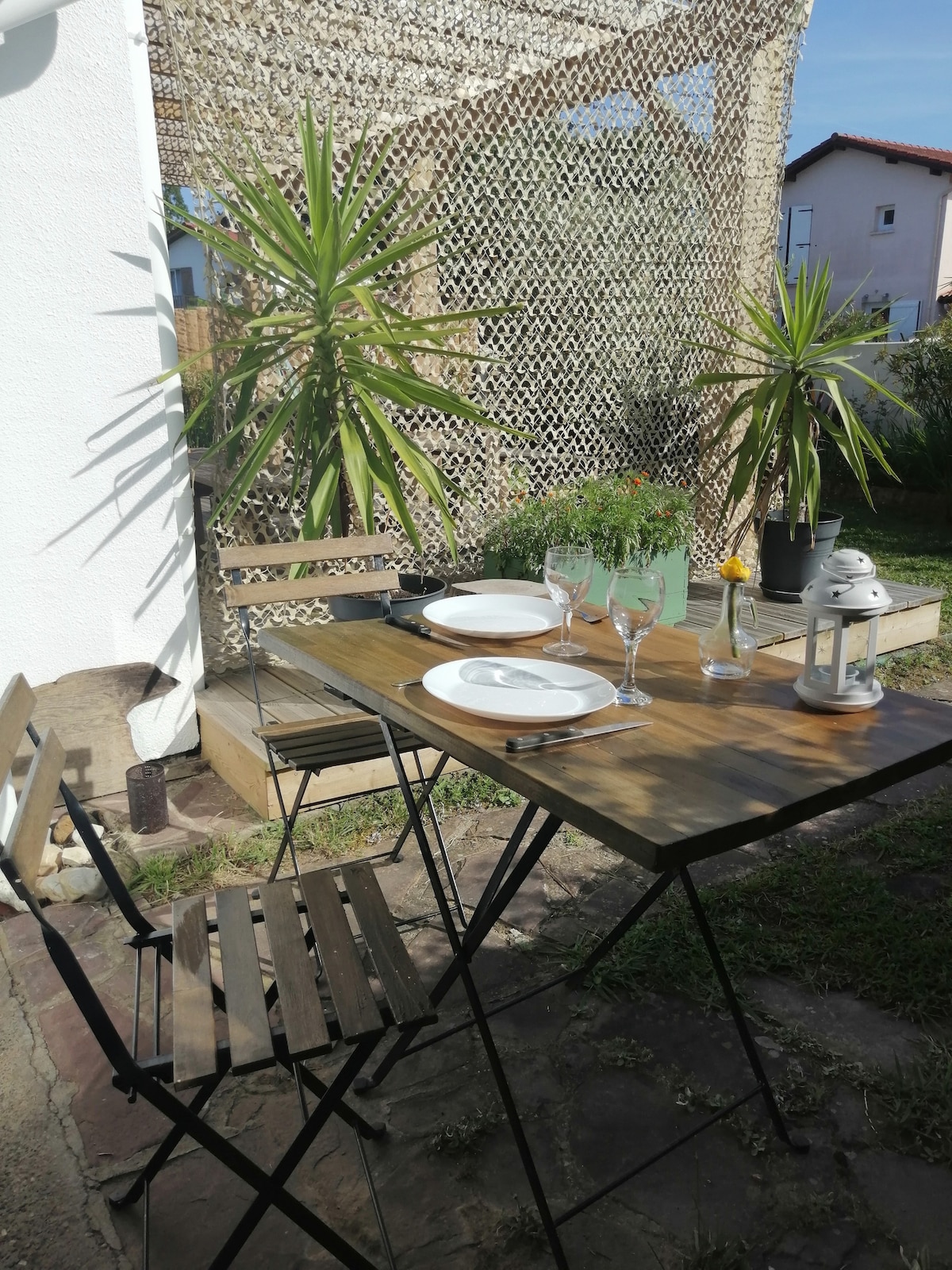 An outdoor dining area is set with a wooden table and two folding chairs, positioned under a decorative privacy screen. Two plates, glasses, and a small candle lantern adorn the table, complemented by potted plants providing a natural touch.
