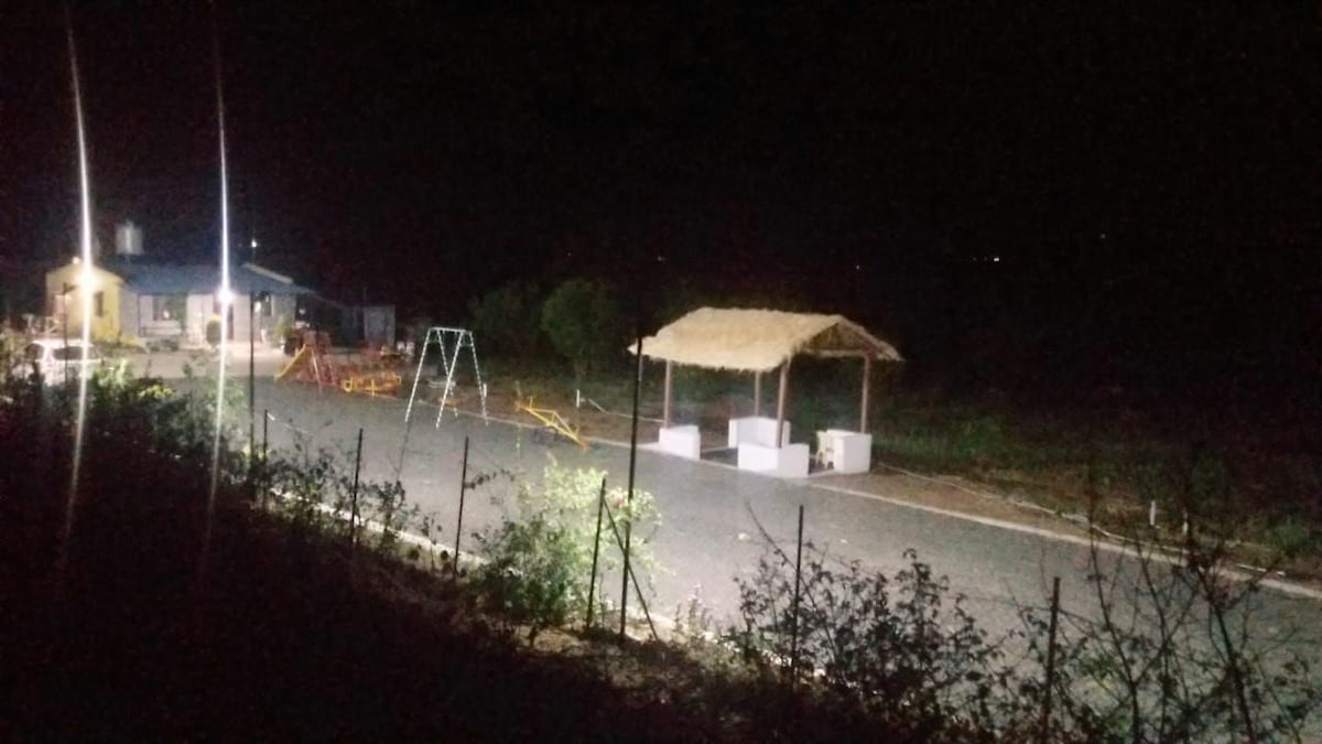 An outdoor area is illuminated at night, featuring a thatched gazebo and a playground with swings. Nearby, a rural building is visible, surrounded by greenery and gravel path. Subtle lighting highlights the tranquil setting, blending nature with recreational facilities.
