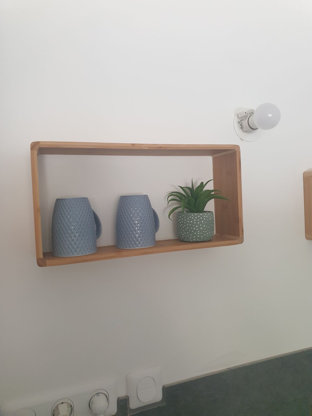 A wooden shelf is positioned against a white wall, displaying two textured blue mugs and a small potted plant. Above, a wall-mounted light fixture emits soft illumination, complementing the subtle decor.