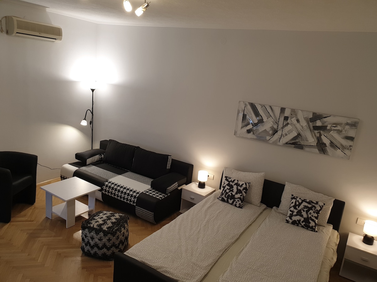 The studio apartment features a bright interior with light-colored walls. Two spacious beds are positioned side by side, covered with patterned bedding. A comfortable black sofa and modern armchair are arranged around a central white coffee table. Soft lighting is provided by floor and table lamps.