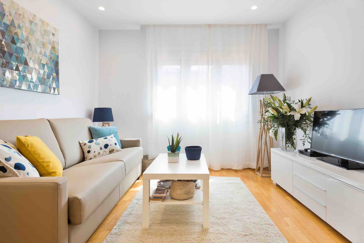 A light-filled living room features a neutral-colored sofa adorned with decorative cushions. A coffee table sits in the center, complemented by a potted plant. A contemporary lamp and a television are positioned nearby, and sheer curtains allow natural light to enhance the space.