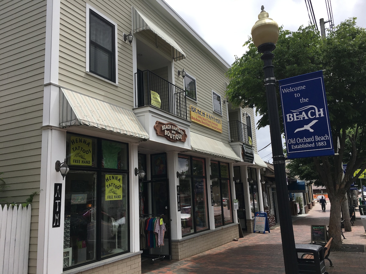 A street view showcases two stories of a commercial building with storefronts below and balconies above. Signs indicate various shops, including a tattoo parlor. A lamppost and a welcome sign for Old Orchard Beach are visible, along with trees lining the walkway.