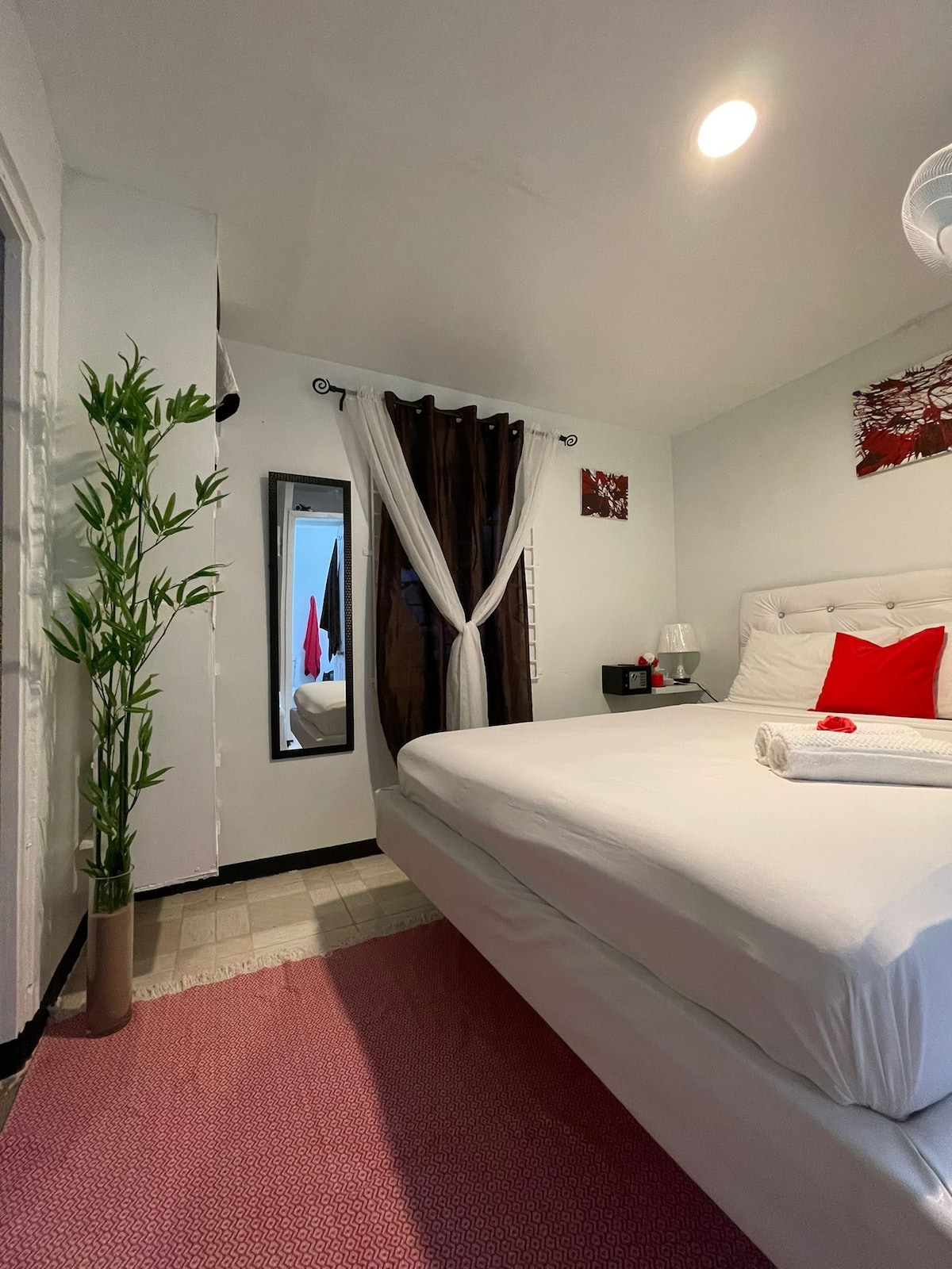 A spacious bedroom is equipped with a modern white bed and a decorative red accent pillow. Soft lighting is provided by overhead fixtures. Dark curtains frame a large mirror, while a decorative plant adds a touch of greenery to the space.