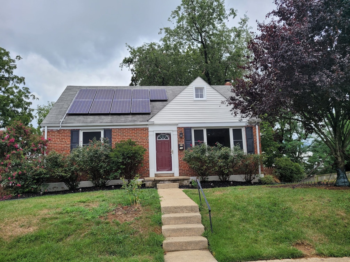 A charming brick house is featured, showcasing solar panels on the sloped roof. The front yard includes manicured shrubs and a walkway leading to the welcoming entryway. Nearby trees provide some shade, creating a tranquil setting in a well-maintained neighborhood.