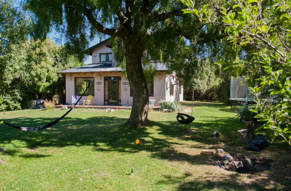 A spacious house is set within a lush, green garden, featuring tall trees providing shade. A hammock is visible under the tree, while a trampoline is located in the background. The exterior showcases large windows that invite natural light into the home.