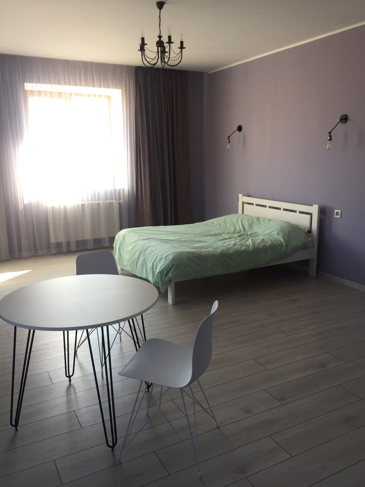 A spacious room features a large bed with a light green blanket, complemented by a simple white headboard. A round table with three chairs is arranged nearby. Natural light enters through a large window, enhancing the lavender walls and creating a calm environment.