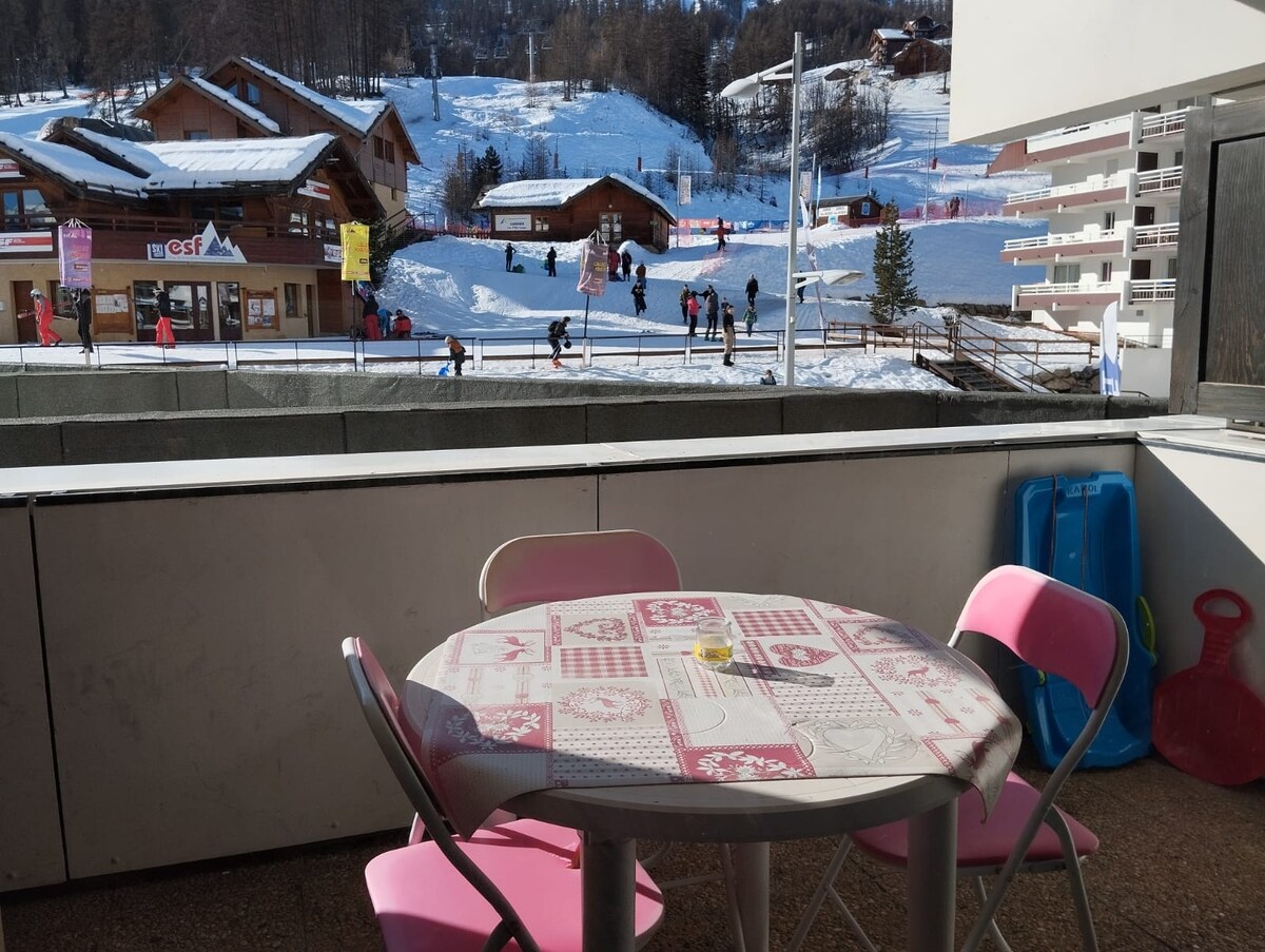 A small balcony is furnished with a round table and two pink folding chairs. The view features snow-covered slopes and wooden chalets, with people enjoying winter activities in the distance. Sunlight brightens the scene, creating a welcoming outdoor space.