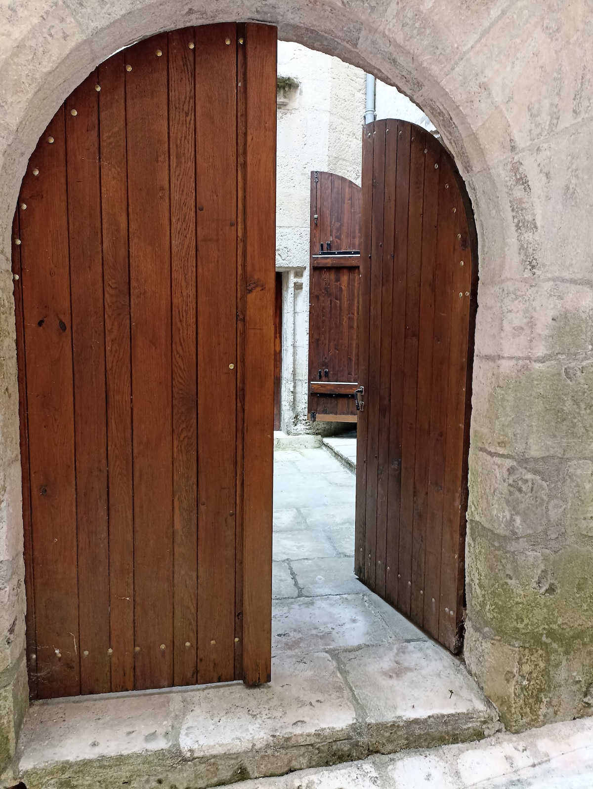 A wooden double door stands ajar, framed by a weathered stone archway. The door, featuring vertical wooden planks, opens into a quaint courtyard paved with stone tiles. Sunlight casts soft shadows on the ground, enhancing the serene ambience of the entrance.