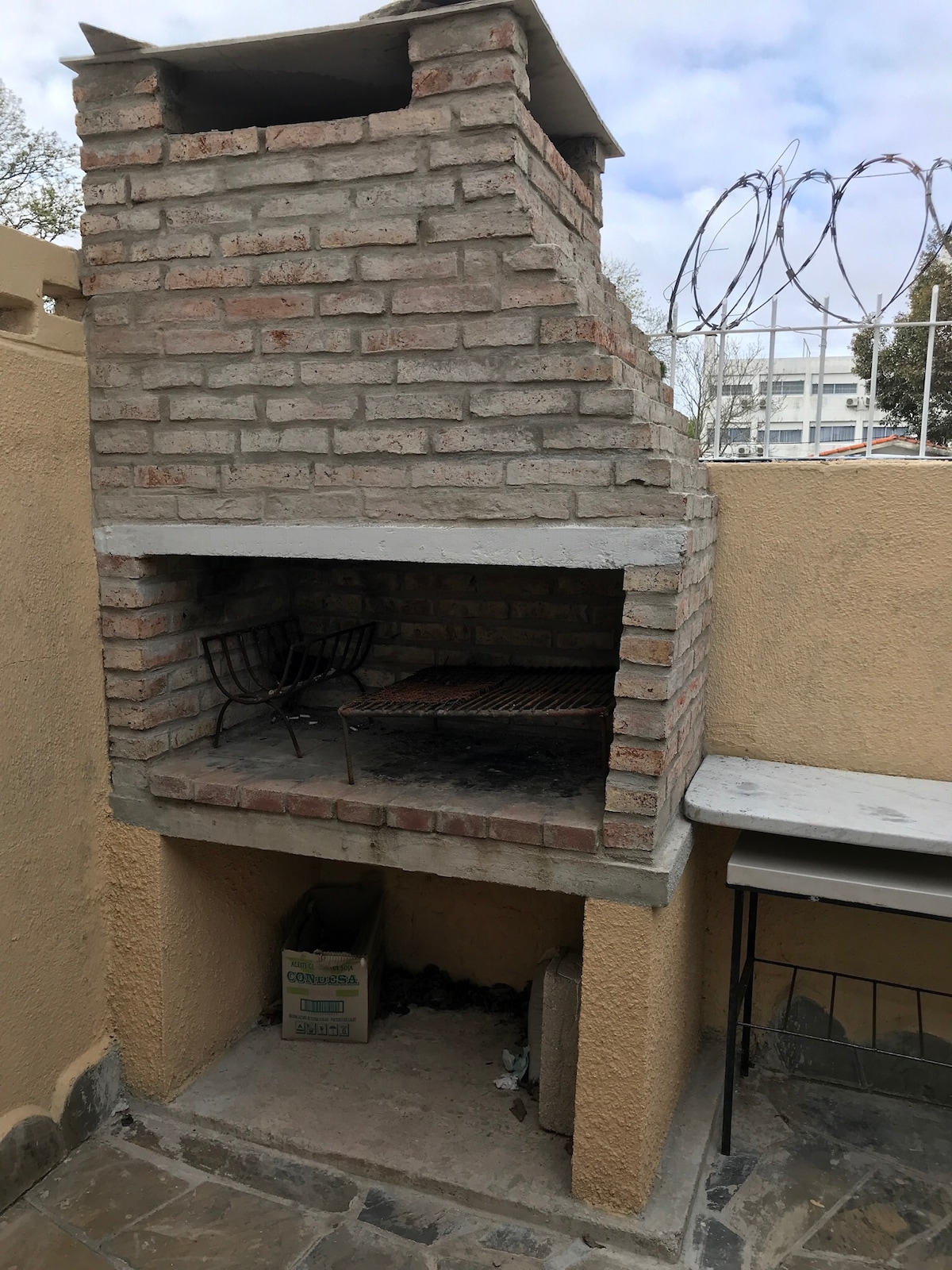 A brick barbecue grill is built into an outdoor wall, featuring a spacious cooking area with a metal grill. The surrounding area includes a small table and a box, with a textured stone surface underneath. A wire fence is visible in the background.