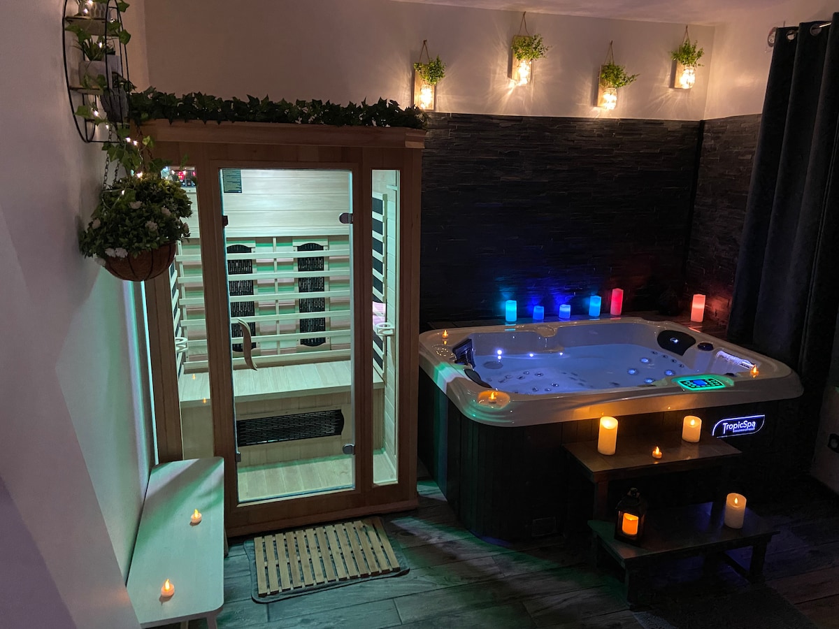 An inviting wellness area features a Jacuzzi with colored lights and a sauna in the background. Soft lighting from wall candles and planters creates a calming ambiance, while a wooden mat lies in front of the Jacuzzi. The space is designed for relaxation.