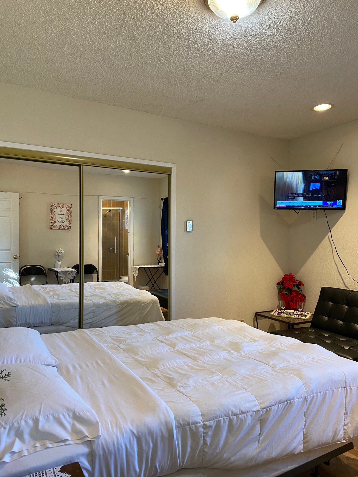 A bright bedroom is presented with a spacious bed covered in white linens. A wall-mounted television displays a news channel. Mirrored closet doors reflect the room, while additional seating is provided by a black sofa. Soft lighting is offered by a ceiling fixture.