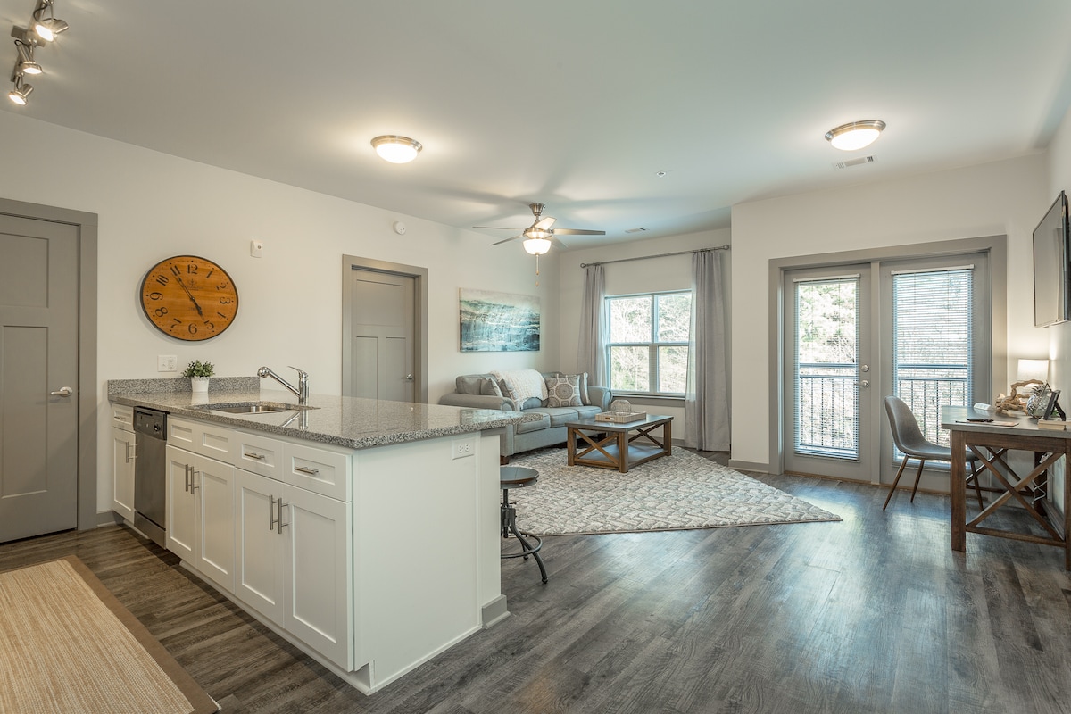 A modern open-concept living area showcases a kitchen with granite countertops and stainless steel appliances. A comfortable seating area is furnished with a light gray sofa and a patterned area rug, while large windows provide natural light and views of the balcony.