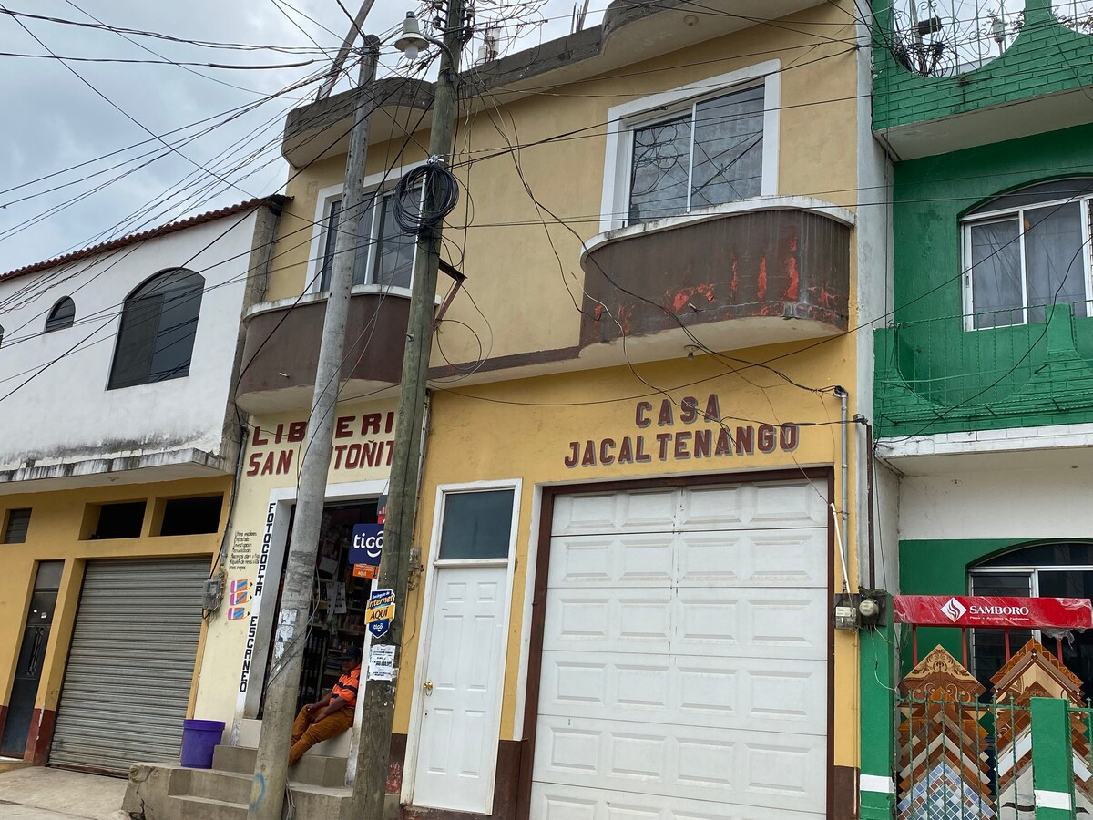 A two-story building is visible, with a yellow facade and a white garage door marked 'CASA JACALTENANGO.' The ground level features a bookshop with signage above its entrance. The second level displays rounded balconies and is slightly weathered, complemented by power lines in the foreground.
