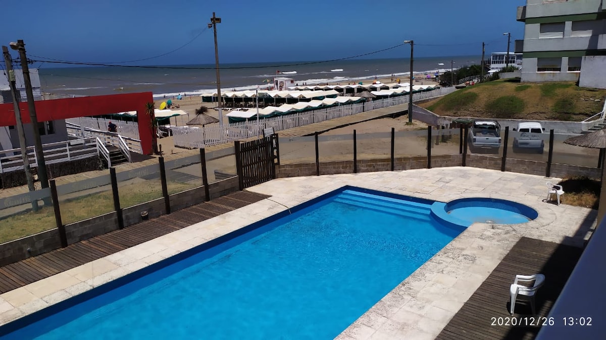 A clear view of a modern outdoor pool is presented, bordered by a wooden deck. The pool's bright blue water reflects the sky. In the background, a beach with umbrellas and lounge chairs can be seen, along with ocean waves rolling onto the shore.