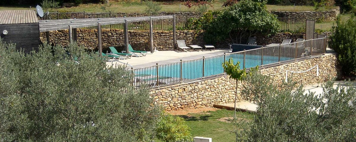 A tranquil outdoor area features a rectangular swimming pool surrounded by a stone wall, with several lounge chairs positioned along the edges. Lush greenery and trees are visible in the background, enhancing the serene setting.