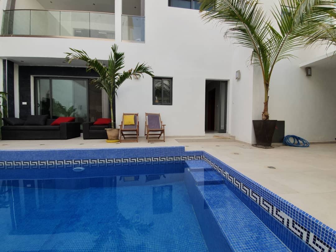 An inviting outdoor area features a shimmering pool with a blue mosaic border. Comfortable seating arrangements are positioned nearby, complemented by tall, leafy plants. The modern villa’s large glass doors provide a view of the interior, creating a seamless connection between spaces.