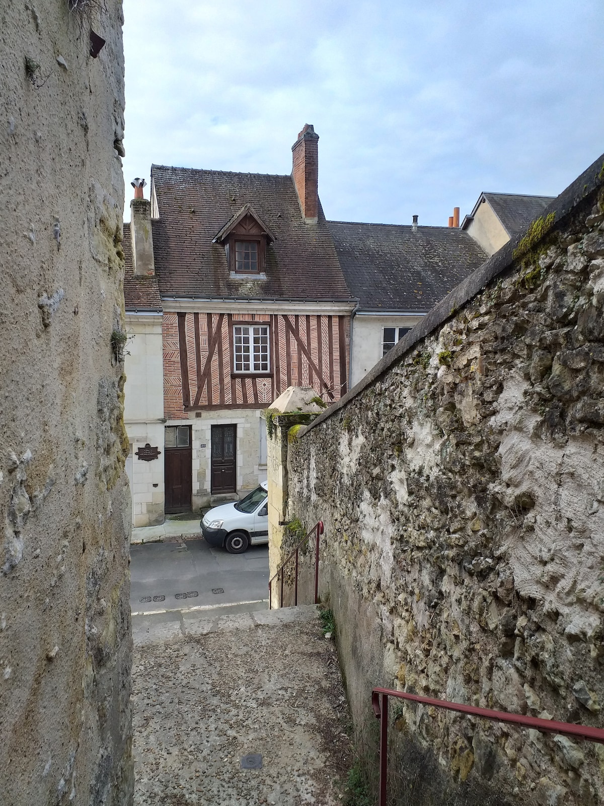 A narrow staircase descends between stone walls, leading to a charming street view. The facade of a historic building features exposed wooden beams and a steep roof, complementing the surrounding architecture. A parked car is visible on the street below.