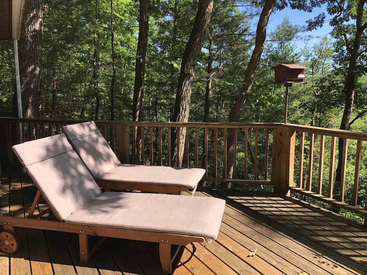 Two lounge chairs with neutral cushions are positioned on a wooden deck, surrounded by lush greenery. Sunlight filters through the trees, casting shadows on the deck surface, creating a relaxing outdoor space.
