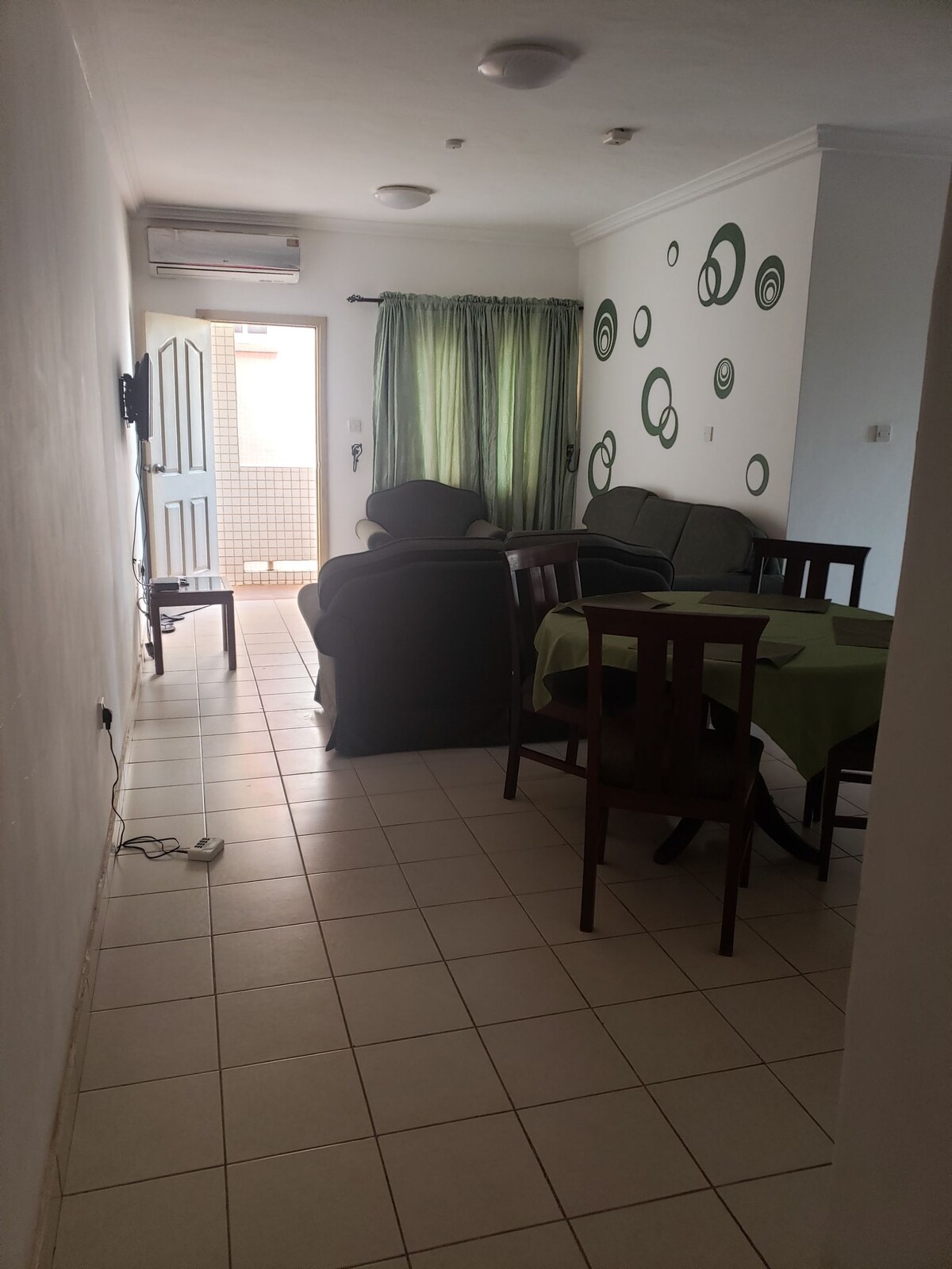 A bright living area is featured, with a green sofa set and a wooden dining table surrounded by chairs. Natural light enters through a door leading to an outdoor space, while textured green wall decorations add interest to the clean, white walls.