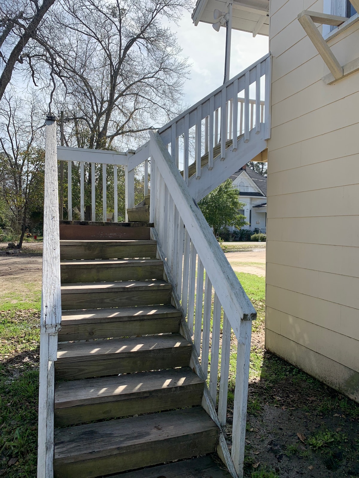 An outdoor staircase is presented, featuring wooden steps and a white railing. The stairs lead to a second-floor entrance, surrounded by grassy areas and trees. A glimpse of the neighborhood can be seen in the background, offering a sense of the tranquil environment.