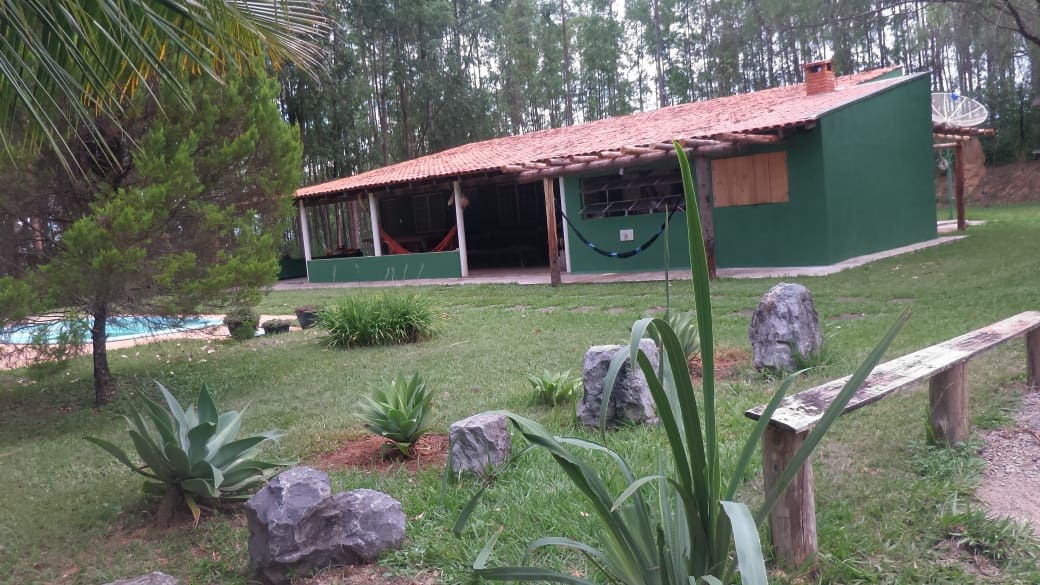 A spacious outdoor area features a green building with a traditional clay tile roof. Hammocks are seen hanging under the porch. A landscaped yard includes decorative rocks and lush plants, with a glimpse of a pool in the background, surrounded by greenery.