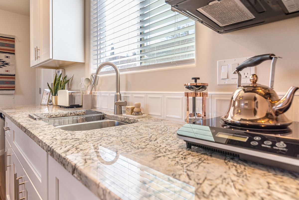 The kitchen area features a polished granite countertop with dual sinks and a modern stainless-steel faucet. A sleek kettle sits beside a selection of kitchen appliances, including an induction cooktop and a stylish coffee maker. Natural light filters in through the nearby window.